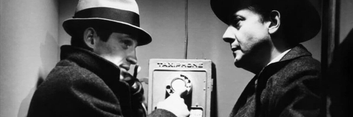 Dwóch mężczyzn w kapeluszach i płaszczach przed automatem telefonicznym