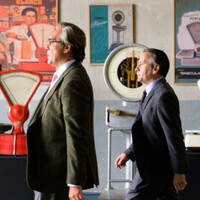 Dwóch starszych mężczyzn idzie korytarzem, w tle stare wagi i plakaty reklamujące wagi 