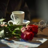 Biała filiżanka stojąca na stole, a obok okulary i dwie czerwone róże 
