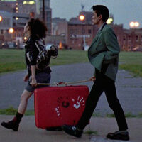 Azjatycka kobieta i mężczyzna idą chodnikiem i niosą wspólnie na patyku czerwoną walizkę