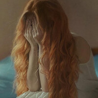 Rudowłosa kobieta ukrywająca twarz w dłoniach, siedząca na łóżku 