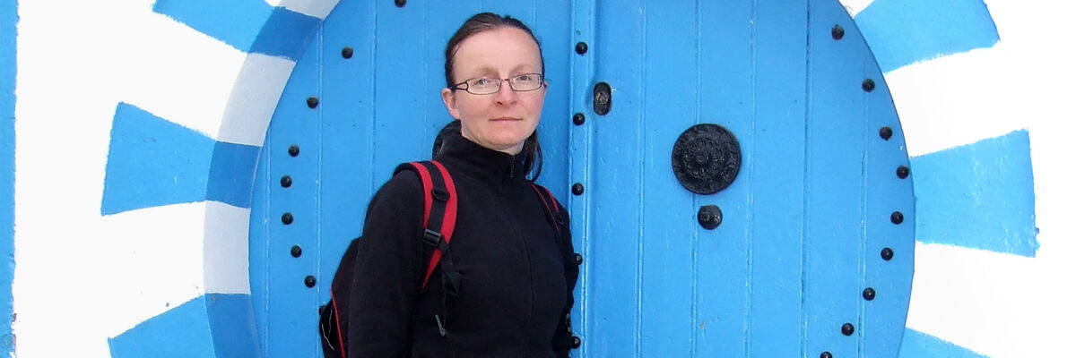 Podróżniczka Ewa Ćwiklińska stojąca w okularach, związanych włosach w czarnej bluzie i plecaku na tle niebieskich drzwi 