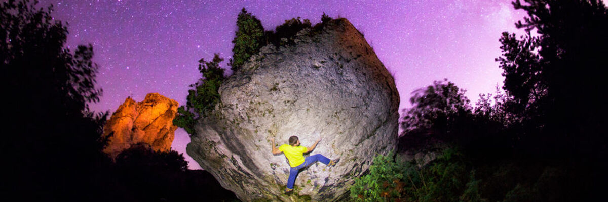 Wspinający się na okrągłą skałę mężczyzna w żółtej koszulce. Niebo jest fioletowe, a wokół znajdują się drzewa