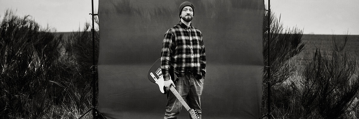 Gitarzysta i wokalista Jakub Sołtysik stojący z gitarą w koszuli w kratę i czapce