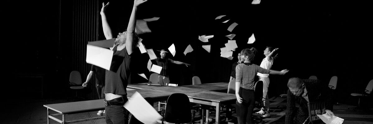 Aktorzy rozrzucający kartki papieru wśród biurek w czarnym pomieszczeniu