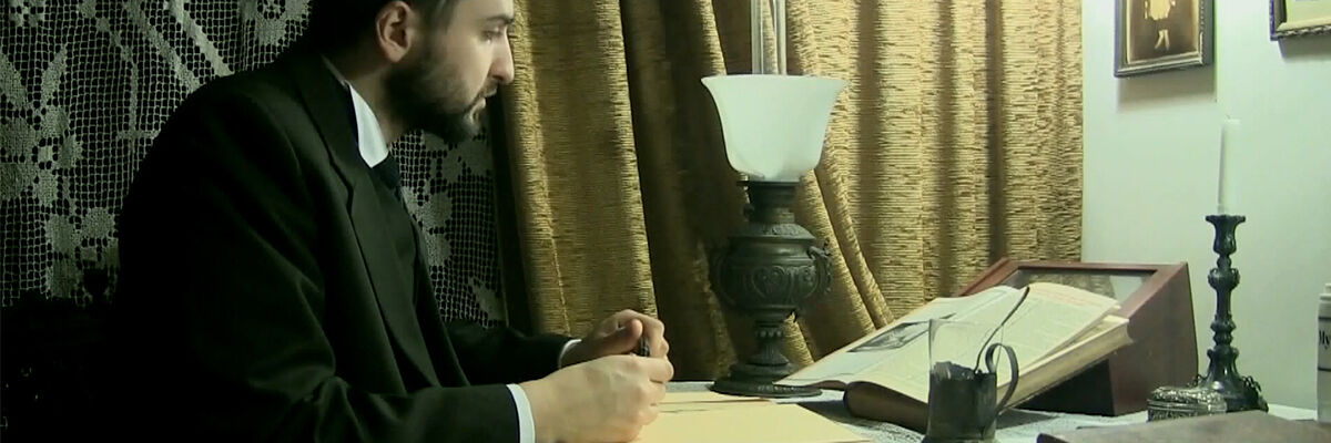 Aktor siedzący za biurkiem z długopisem w ręce, a przed nim otwarta książka i szklanka po prawej stronie i lampka po lewej stronie