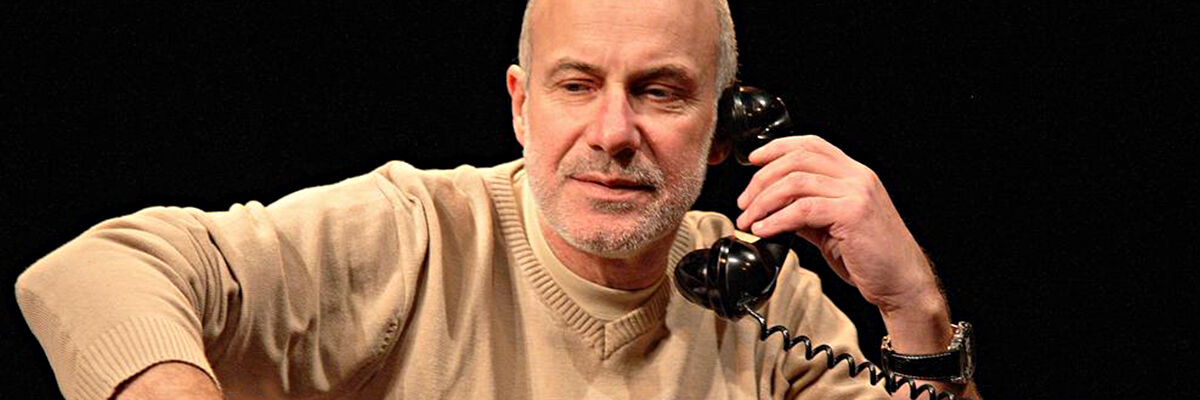 Piotr Machalica w beżowej koszuli trzymający słuchawkę czarnego telefonu na czarnym tle