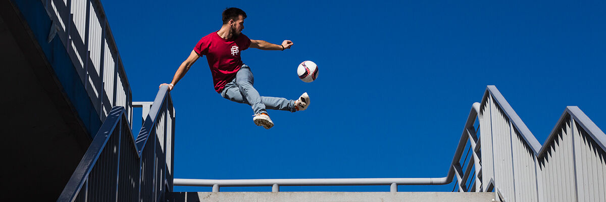 Chłopak w czerwonej koszulce trzymający się za obręcze schodów i odbijający nogą piłkę podczas skoku 