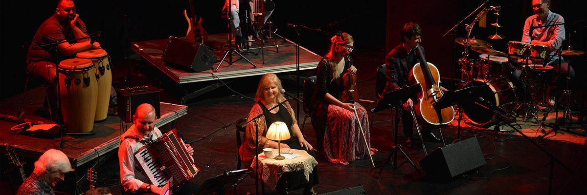 Aktorka Stanisława Celińska siedząca na scenie razem z artystami grającymi na akordeonie, bębnach, gitarze i perkusji