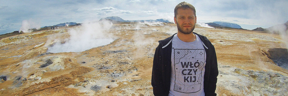Podróżnik Przemysław Rychter stojący w bluzie i koszulce z napisem "Włóczykij" na tle wulkanu