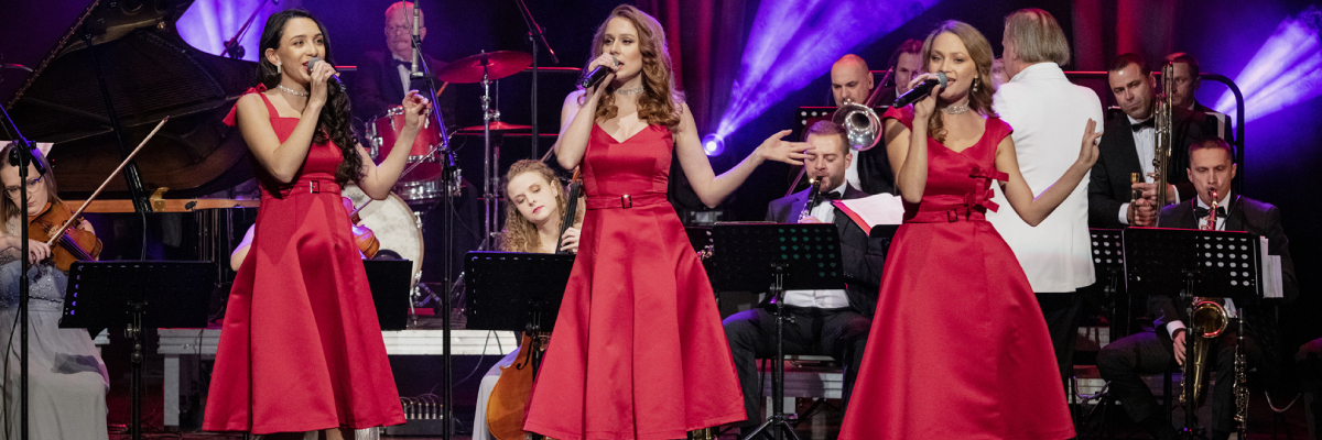 Stojące na scenie trzy wokalistki w czerwonych sukniach trzymające mikrofony w prawej ręce