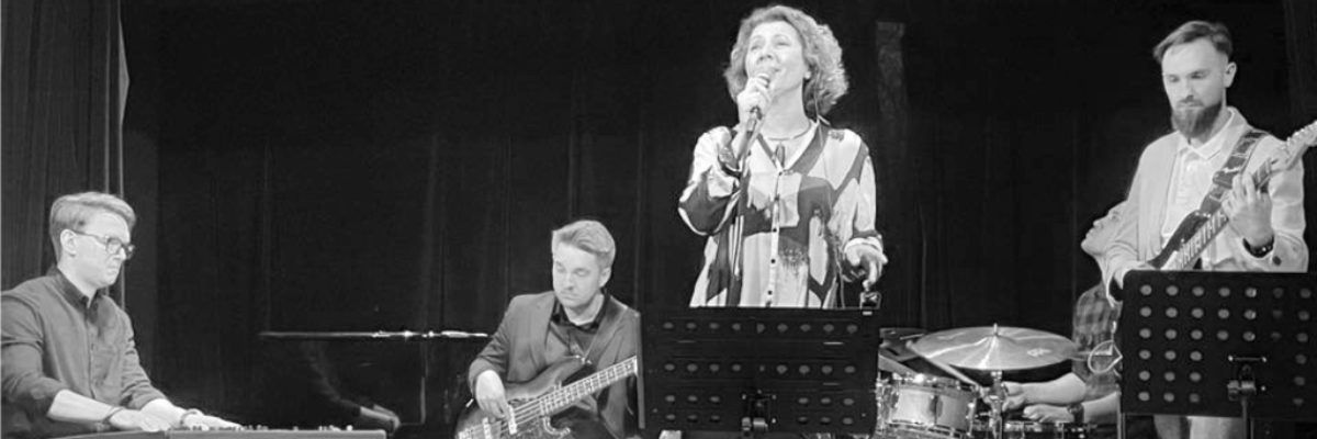 Kobieta śpiewająca na scenie, a obok dwóch grających na gitarze mężczyzn i jeden na keyboardzie