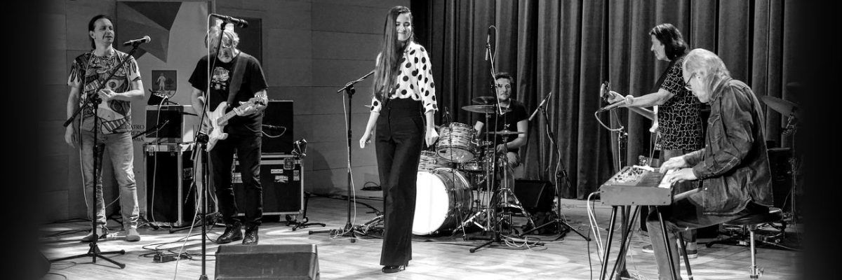 Kobieta w czarnych spodniach i białej bluzce w czarne kropki stojąca przed mikrofonem, a obok niej muzycy za nią muzycy. 