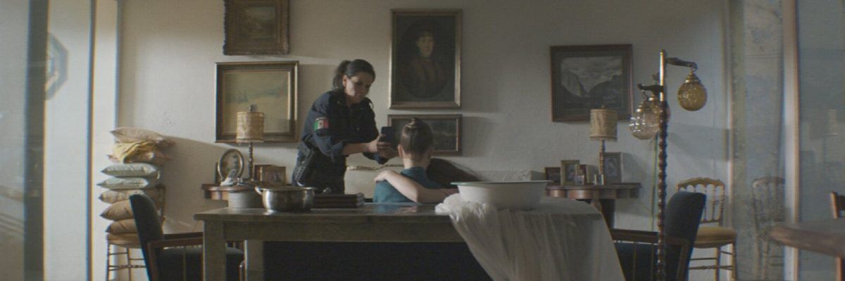 Kadr z filmu "Ukryty klejnot" gdzie kobieta trzyma w ręce telefon komórkowy siedzącej na krześle kobiecie, którą przytula inna kobieta. Za nimi stoi biurko, na którym jest biała miska, a pod nią biała tkanina