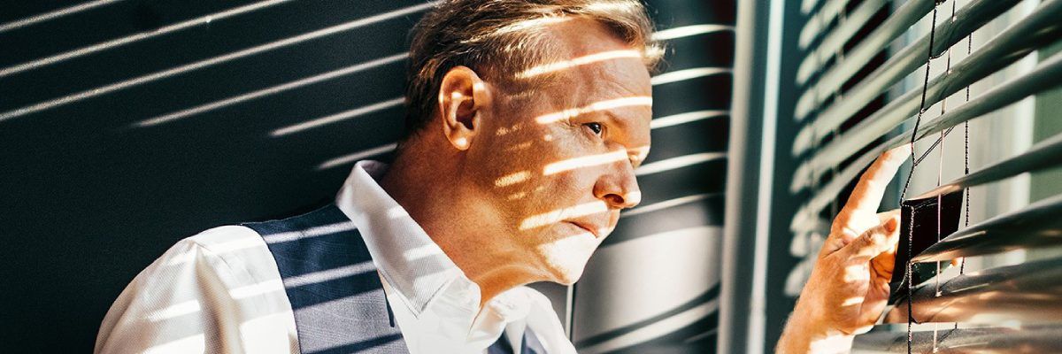 Michał Bajor w białej koszuli patrzący się przez okno, na których są żaluzje, przez które pada światło na jego twarz.