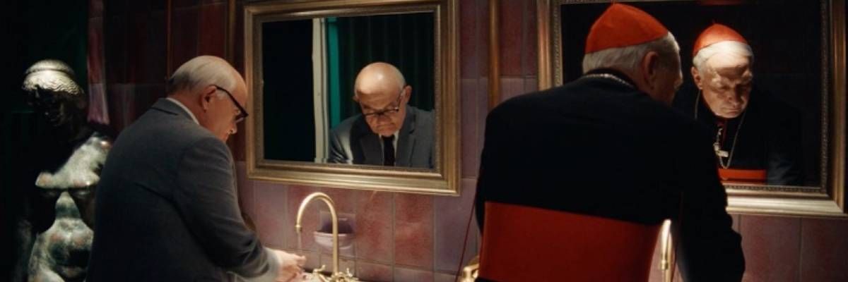 Stojący w garniturze przed lustrem i umywalką mężczyzna, a obok niego również stojący arcybiskup przed lustrem