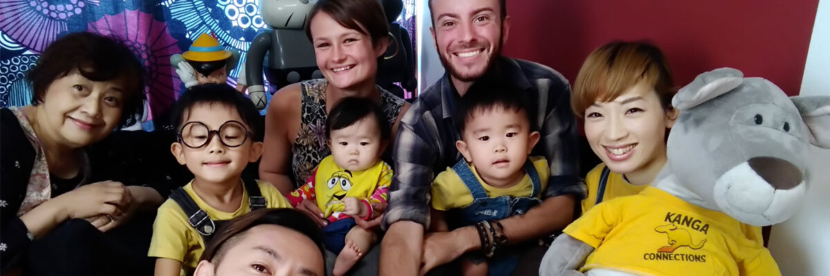 Podróżniczka pozująca do zdjęcia z mężczyzną obok, starszą panią, trzema azjatyckimi dziećmi oraz panią trzymającą pluszowego kangura ubranego w żółtą bluzkę