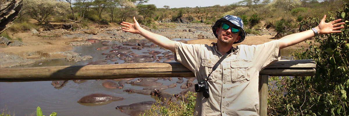 Podróżnik Robert Garus z otwartymi rękami w kapeluszu i okularach słonecznych i przewieszonym przez ramie aparatem fotograficznym, a w tle hipopotamy w wodzie