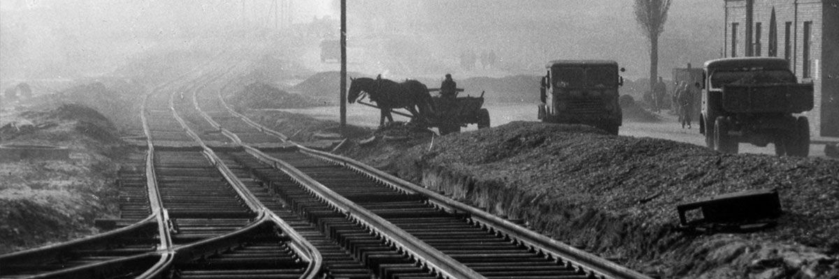 Czarno-biała fotografia torów tramwajowych i powozu konnego obok dwóch samochodów z końca lat dwudziestych XX w.
